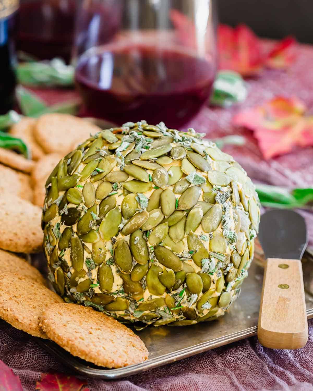 A large cheese ball coated in pumpkin seeds is displayed on a tray with crackers and a butter knife, with a glass of red wine in the background.