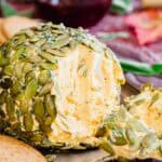 A close-up of a pumpkin seed-coated cheese ball on a tray with a wedge cut out, showing its creamy interior. Crackers and a glass of red wine are visible in the background.