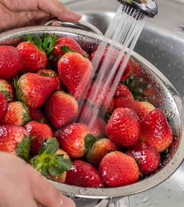 A person rinses fresh strawberries with water in a metal colander over a kitchen sink.