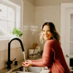 Sara Nelson, wearing a red sweater, smiles while washing something under a faucet in her bright kitchen.