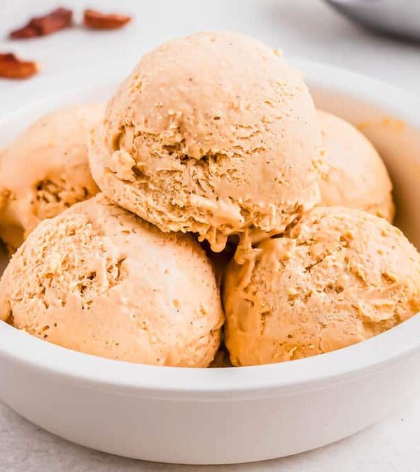A white bowl contains five scoops of sweet potato ice cream on a light-colored surface. Nearby are cinnamon sticks, pecans, and spoons.