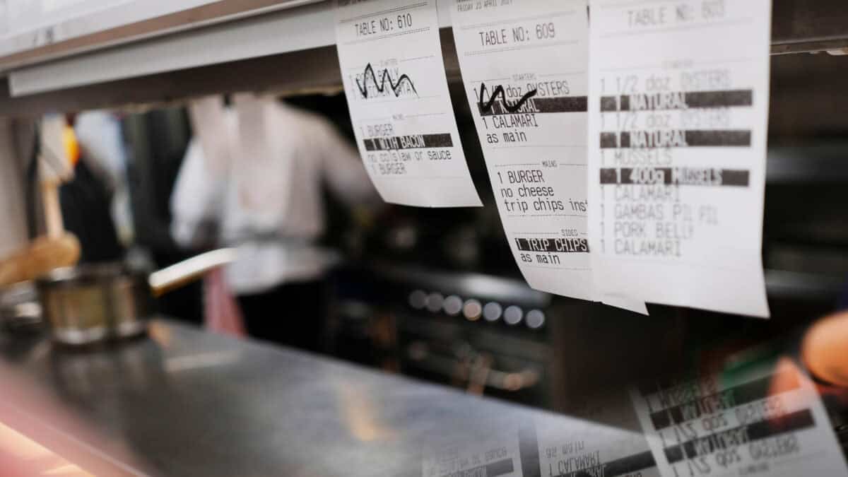 Close-up of several printed restaurant order tickets hanging on a metal rack in a kitchen, with a blurred stove and cooking area in the background.