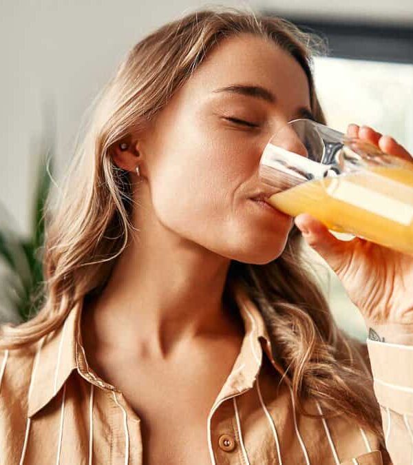 A person with long hair drinks orange juice from a glass while standing indoors near a plant.