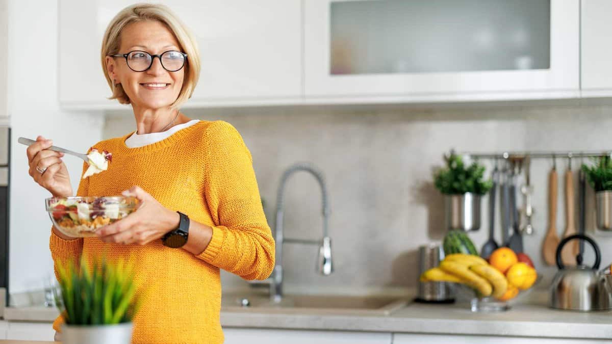 A woman with short blonde hair and glasses is smiling while eating a bowl of salad in a modern kitchen.