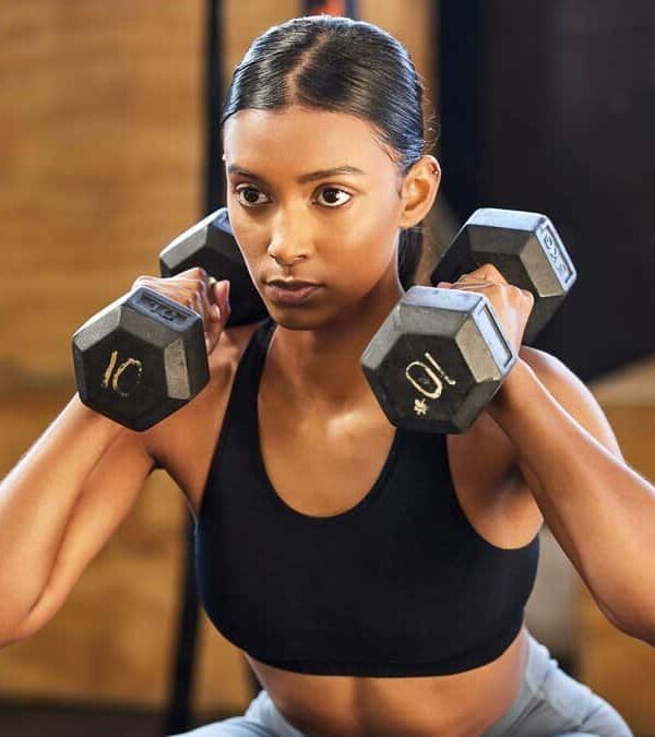 A person in athletic wear performs a squat exercise while holding 10-pound dumbbells on their shoulders, in an indoor gym setting.