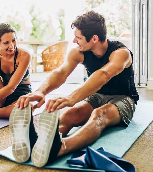 A man and a woman are sitting on yoga mats inside a well-lit room, stretching and smiling at each other. She is tying her shoe; he is reaching for his toes. Exercise equipment is nearby.