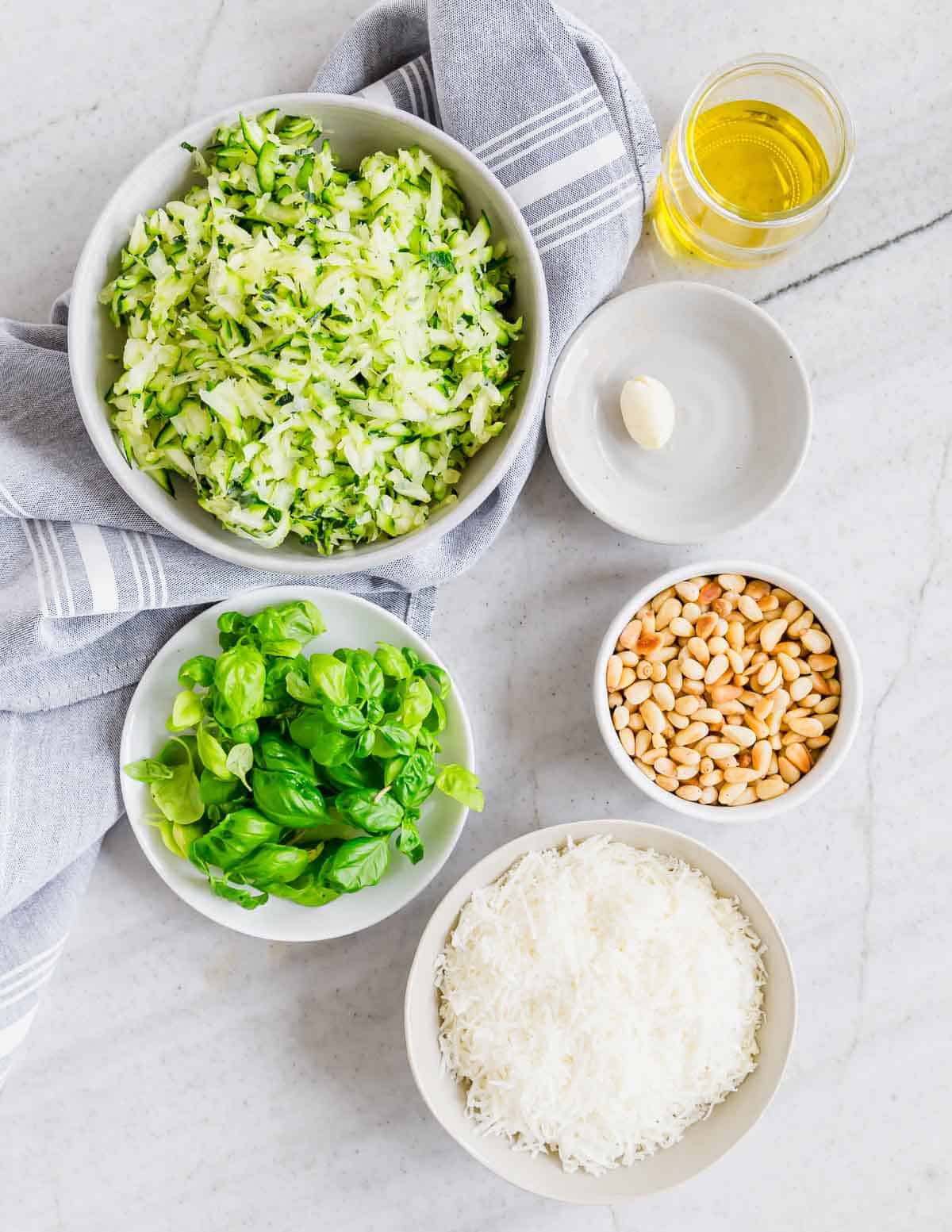 Ingredients for pesto laid out on a marble surface: shredded zucchini, a glass of oil, garlic clove, pine nuts, fresh basil, grated cheese, and a gray-striped napkin.