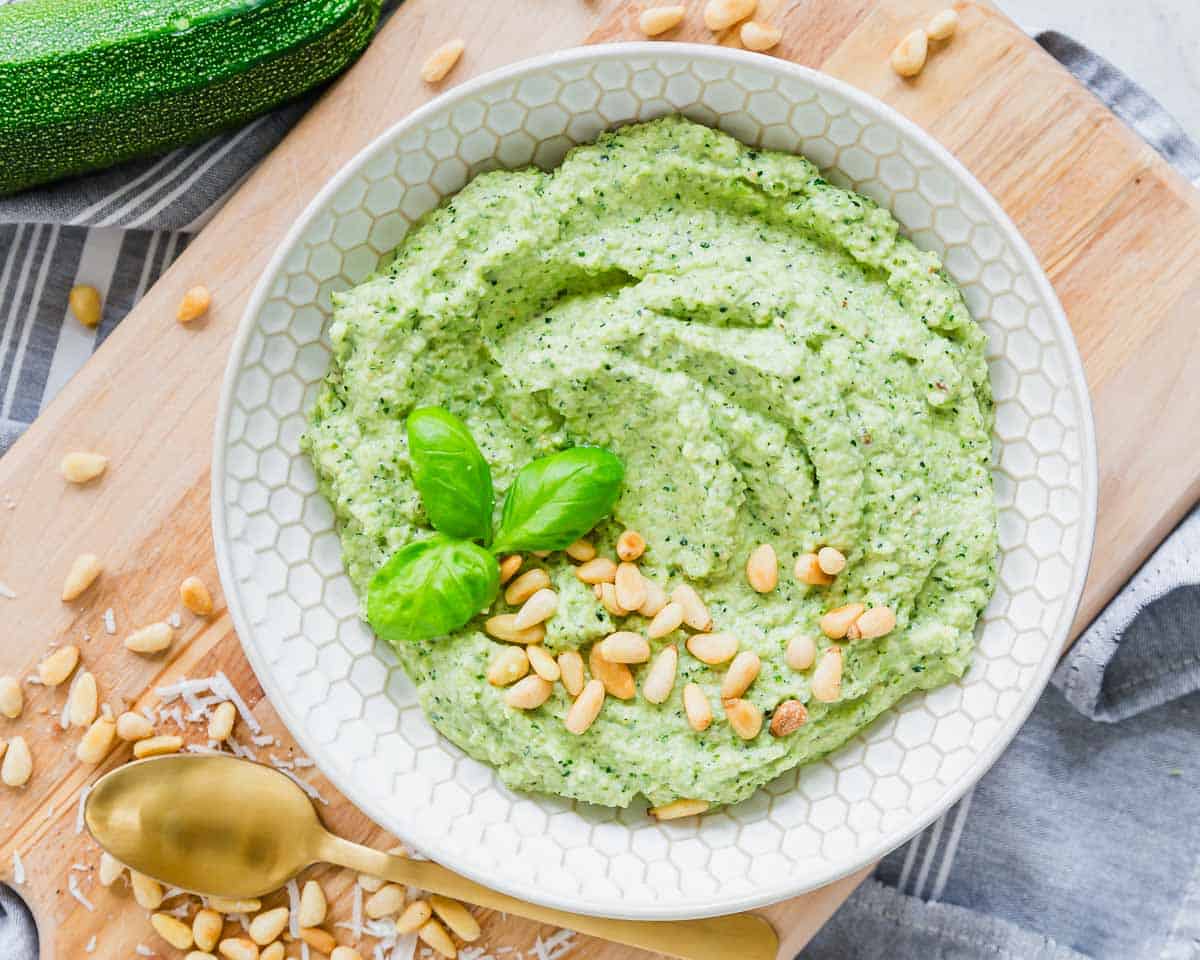 A bowl of zucchini pesto garnished with basil leaves and pine nuts on a wooden board, with a gold spoon nearby.