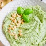 A bowl of pesto made with zucchini is garnished with pine nuts and basil leaves. A gold spoon and some scattered pine nuts are in the background.