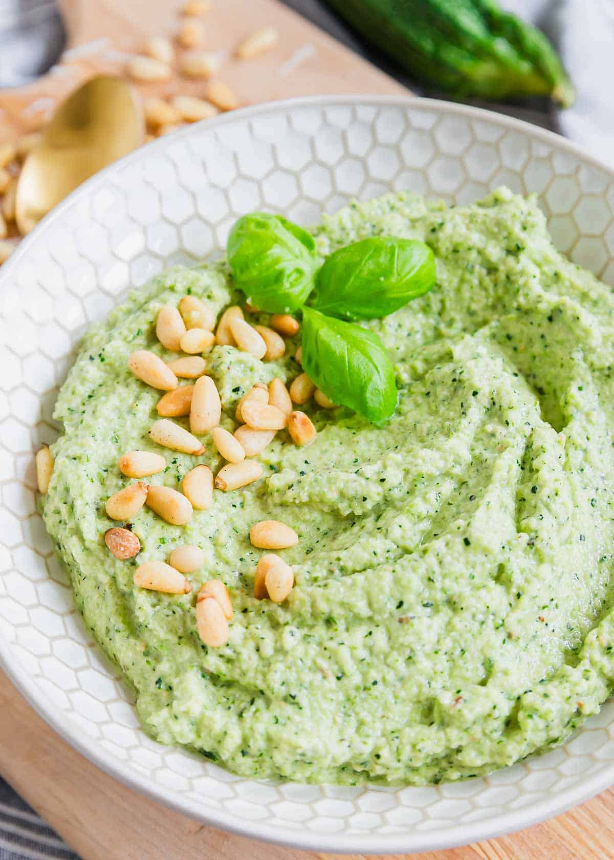 A bowl of pesto made with zucchini is garnished with pine nuts and basil leaves. A gold spoon and some scattered pine nuts are in the background.