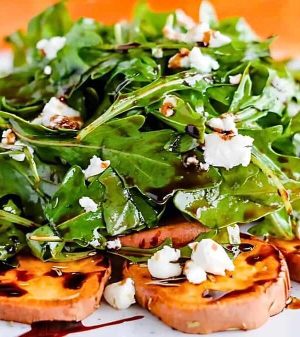 A salad featuring arugula with crumbled cheese on a bed of roasted sweet potato slices, drizzled with balsamic glaze.