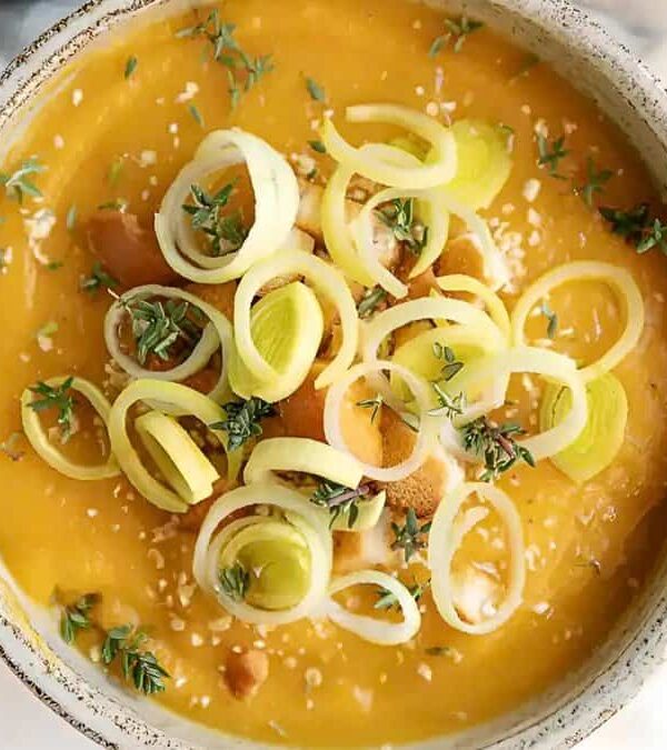 A bowl of creamy pumpkin soup garnished with sesame seeds, sliced leeks, herbs, and bread croutons.