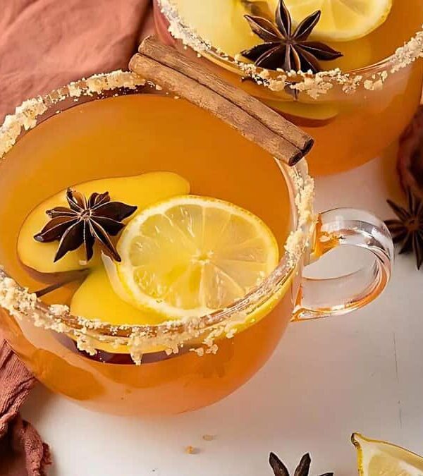 Two glass mugs of apple cider garnished with lemon slices, star anise, and a cinnamon stick sit on a brown cloth. An apple and loose star anise are nearby.