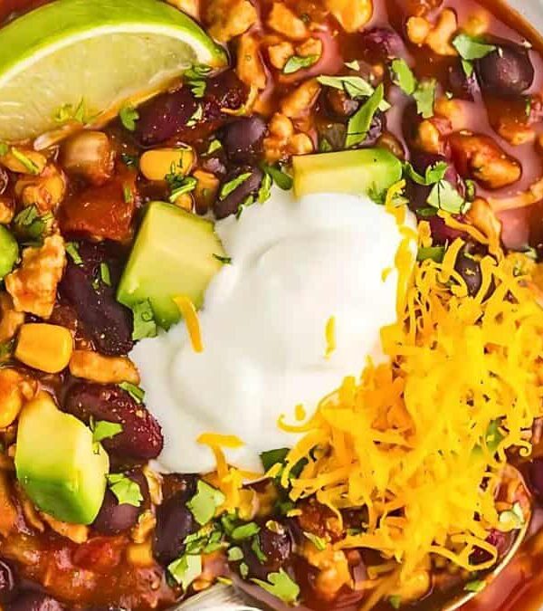 A bowl of chili garnished with sour cream, shredded cheese, avocado chunks, and a lime wedge. The chili contains beans, corn, and ground meat, and is sprinkled with chopped herbs.