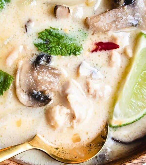 A bowl of creamy soup with mushrooms, red chilies, lime slice, and herbs, served with a gold and black spoon.