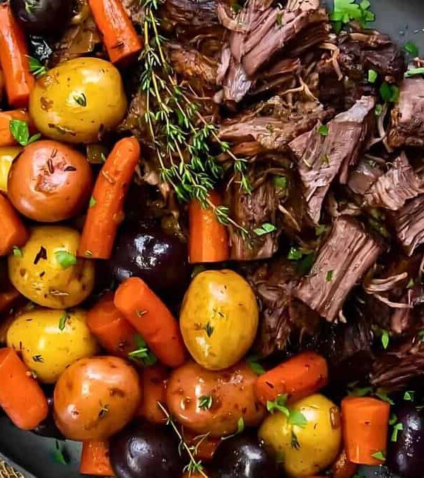 A plate of pot roast with shredded meat, assorted roasted potatoes, carrots, and garnished with fresh parsley and thyme.