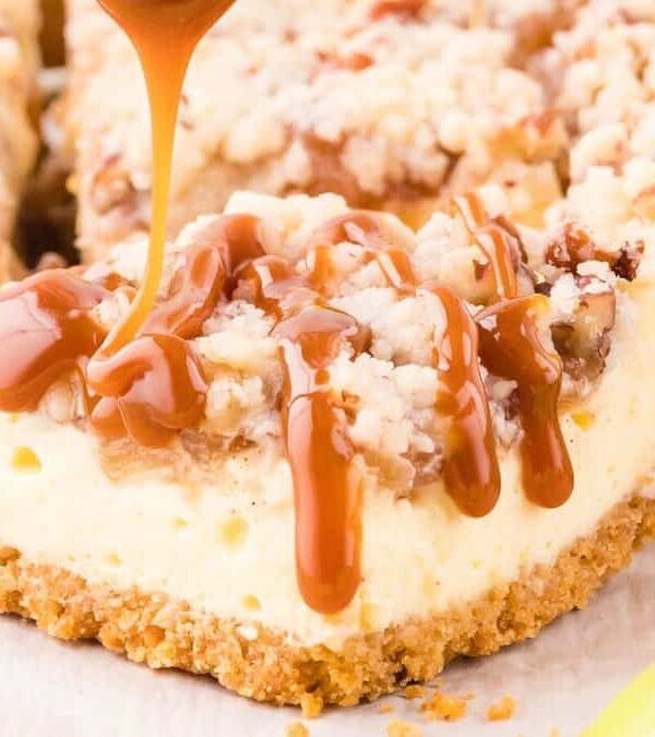 Close-up of a caramel drizzle being poured onto a cheesecake bar with a crumbly topping and a crumb crust. A green apple slice is partially visible in the corner.