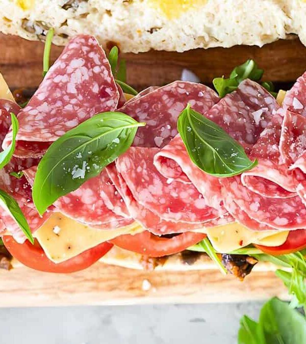 Close-up of an open sandwich with sliced salami, cheese, tomatoes, leafy greens, and basil leaves on a wooden board.