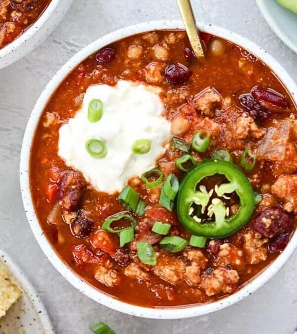 A bowl of chili topped with sour cream, sliced jalapeno, and green onions next to a plate of cornbread and sliced avocado on a light grey surface.