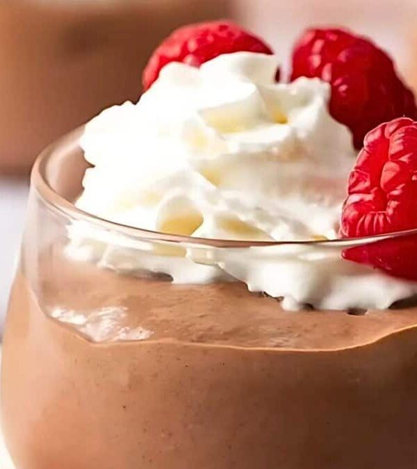 A glass cup filled with chocolate mousse, topped with whipped cream and garnished with three raspberries. Another dish with chocolate mousse and chia seeds can be seen in the background.