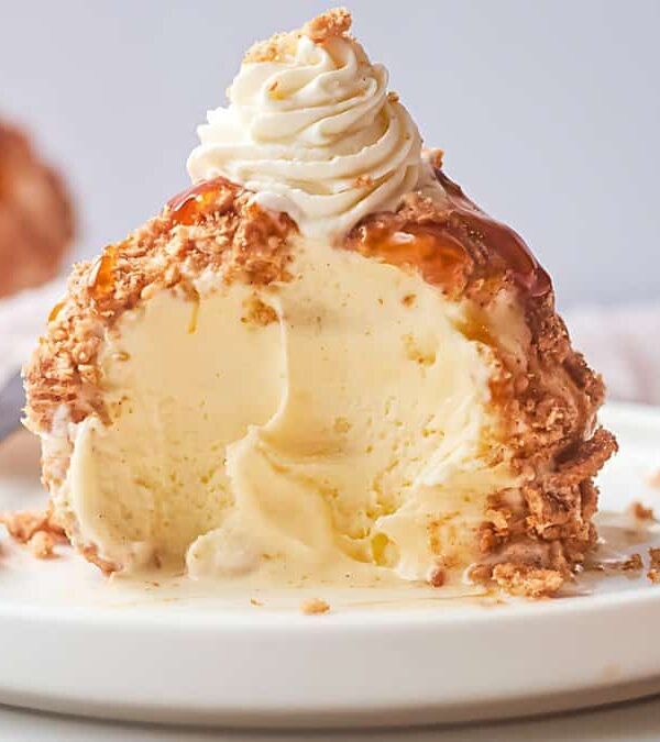 A partially eaten dessert featuring a vanilla ice cream core with a crumbly coating and a dollop of whipped cream on top, served on a white plate. A spoon rests beside the dessert.