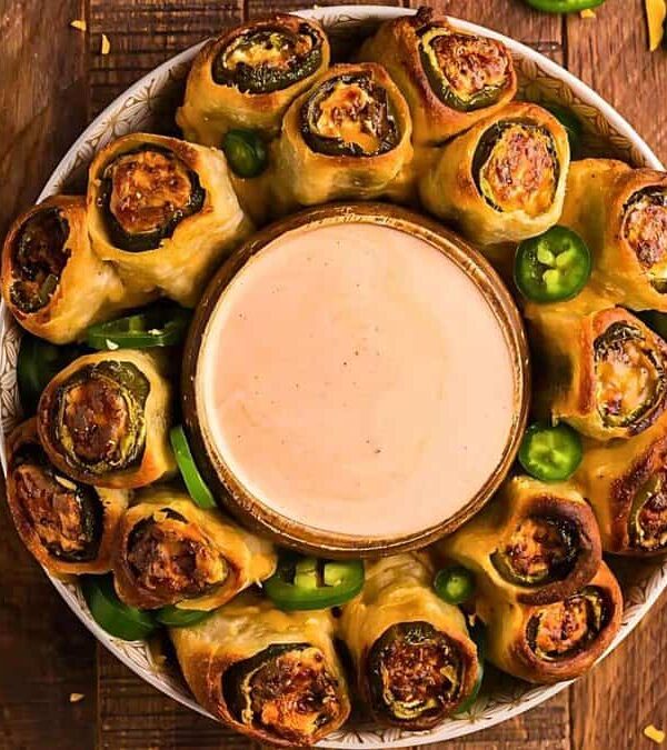A plate of bacon-wrapped jalapeño poppers surrounds a bowl of dipping sauce on a wooden surface, with shredded cheese and whole jalapeños scattered around.