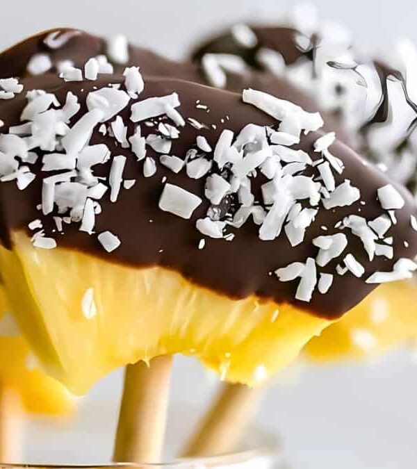 Close-up of chocolate-covered pineapple slices on wooden sticks, with a sprinkling of shredded coconut on top.