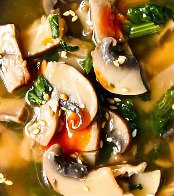 A close-up of a bowl of miso soup containing mushrooms, spinach, and sprinkled with sesame seeds. The blue ceramic bowl has a decorative pattern.