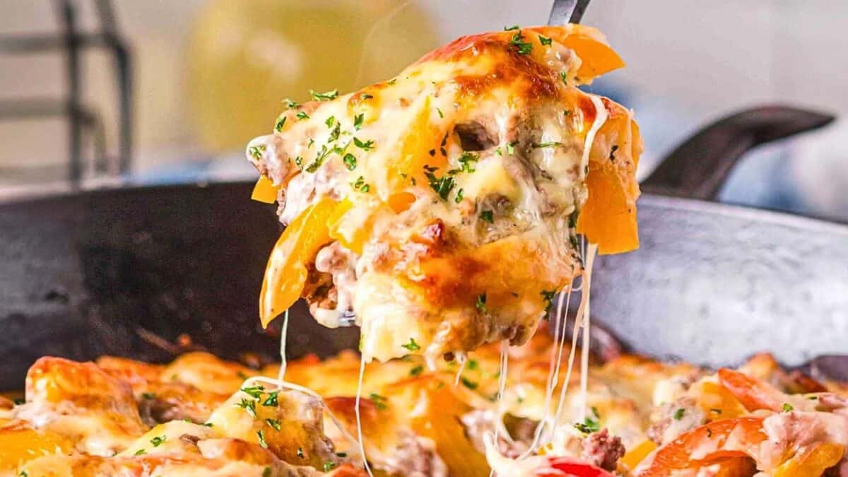 A close-up of a cheesy casserole being lifted from a skillet, showcasing layers of melted cheese, ground meat, bell peppers, and garnished with herbs.