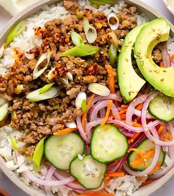A bowl with rice, ground meat, sliced avocado, red onion, cucumber, and shredded carrots garnished with scallions and sesame seeds, with a fork resting on the side.