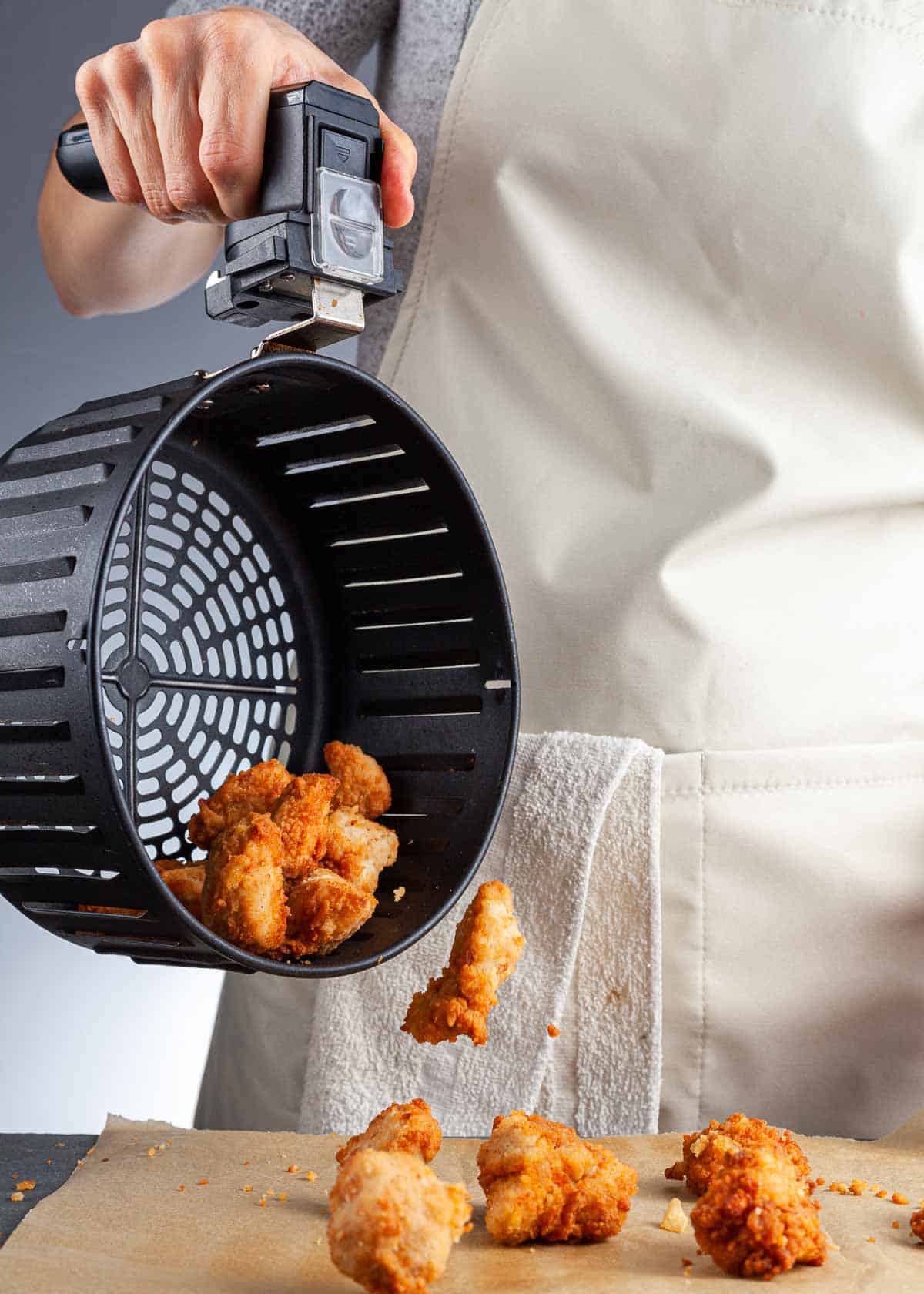 Person pouring fried chicken pieces from an air fryer basket onto parchment paper.