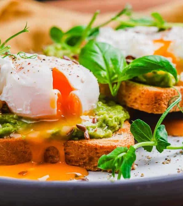 Two slices of toast topped with mashed avocado and poached eggs, garnished with greens and spices, with yolks slightly oozing.