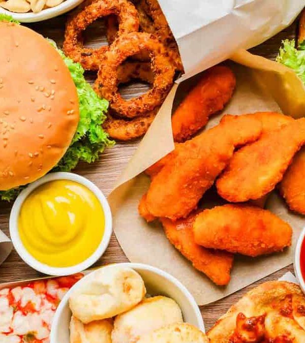 A variety of fast food including burgers, hot dogs, onion rings, chicken tenders, fries, a slice of pizza, and dipping sauces are spread out on a table.
