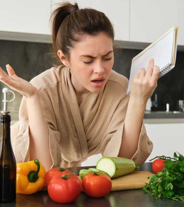 A woman in a kitchen, wearing a robe, holding a notepad, looks frustrated while surrounded by various vegetables including tomatoes, zucchini, bell peppers, and herbs on the counter.