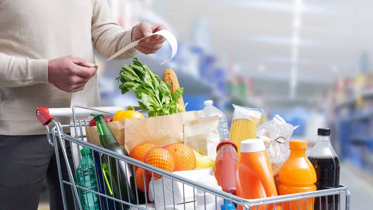 A person holding a receipt stands next to a shopping cart filled with groceries, including vegetables, drinks, and packaged items, in a store aisle.