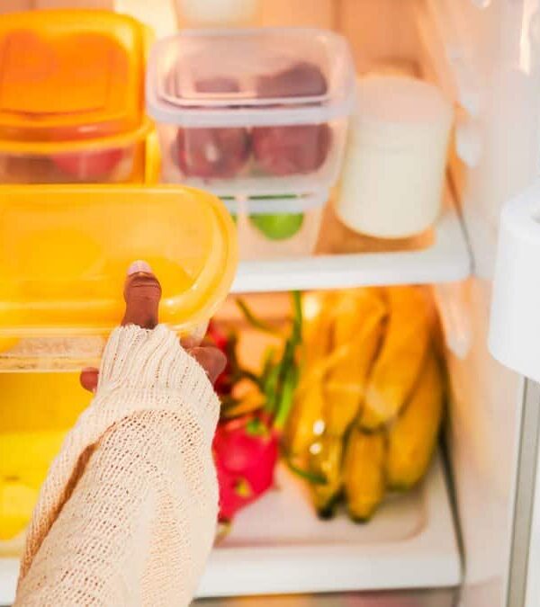 Person placing a yellow-lidded container on the middle shelf of a refrigerator containing various containers and canned items.