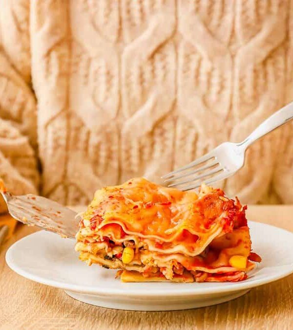 Person in a beige sweater holding a fork and knife, about to eat a piece of lasagna on a white plate.