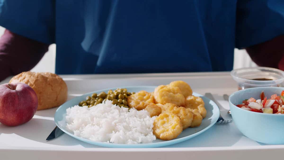 A person's meal tray with a plate containing rice, green peas, and breaded chicken nuggets. The tray also includes an apple, a bread roll, a bowl of salad, and a small cup of sauce.