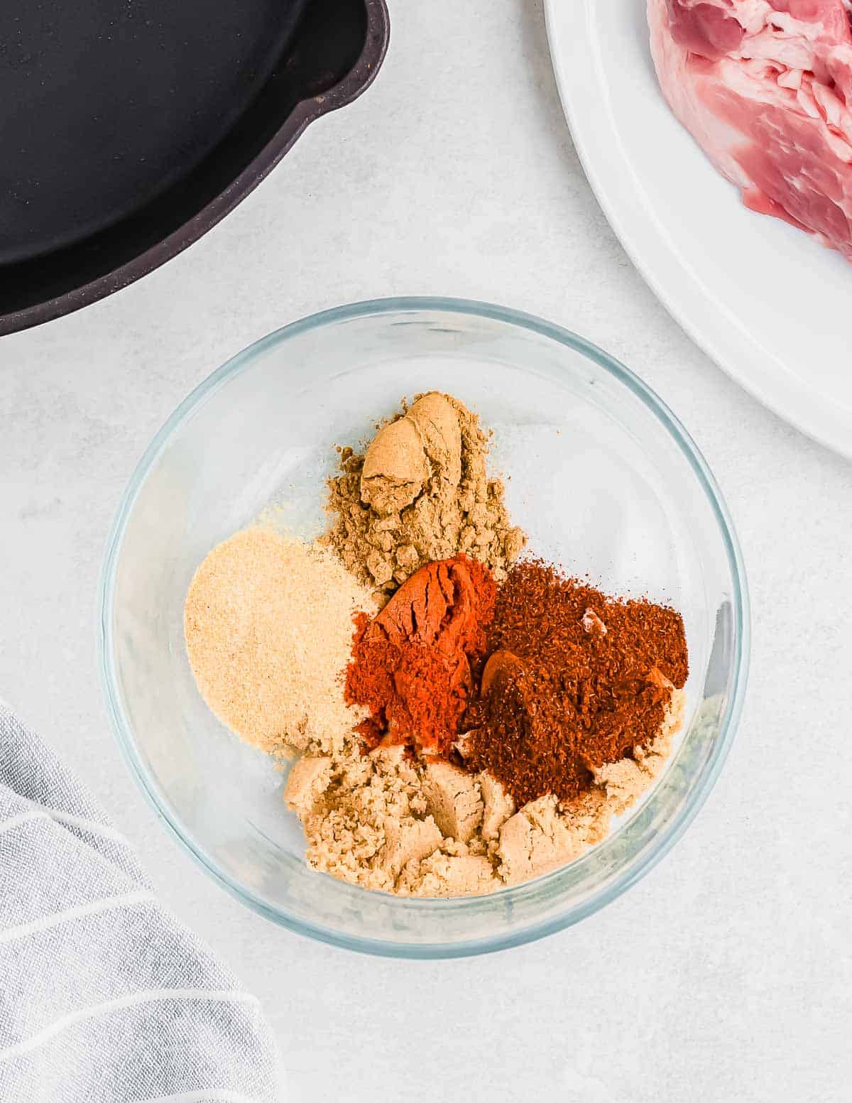 A glass bowl filled with a variety of spices, including paprika, brown sugar, and garlic powder, with a raw piece of meat on a nearby plate and a cast iron pan in the corner.