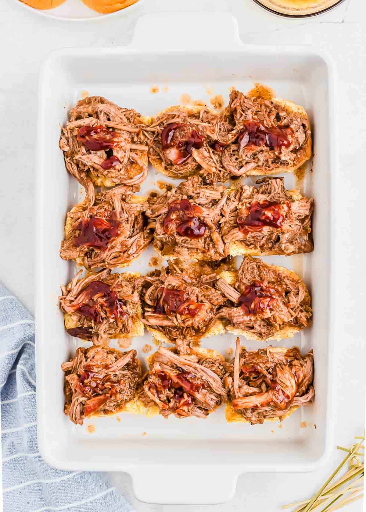 A white baking dish containing twelve bun bottoms topped with pulled pork and barbecue sauce. A blue and white striped cloth is partially visible on the bottom left.