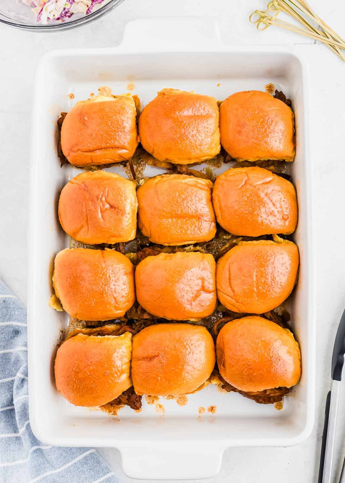A white rectangular baking dish containing twelve evenly spaced pulled pork sliders with golden brown buns. A folded blue and white kitchen towel is partially visible on the left side.