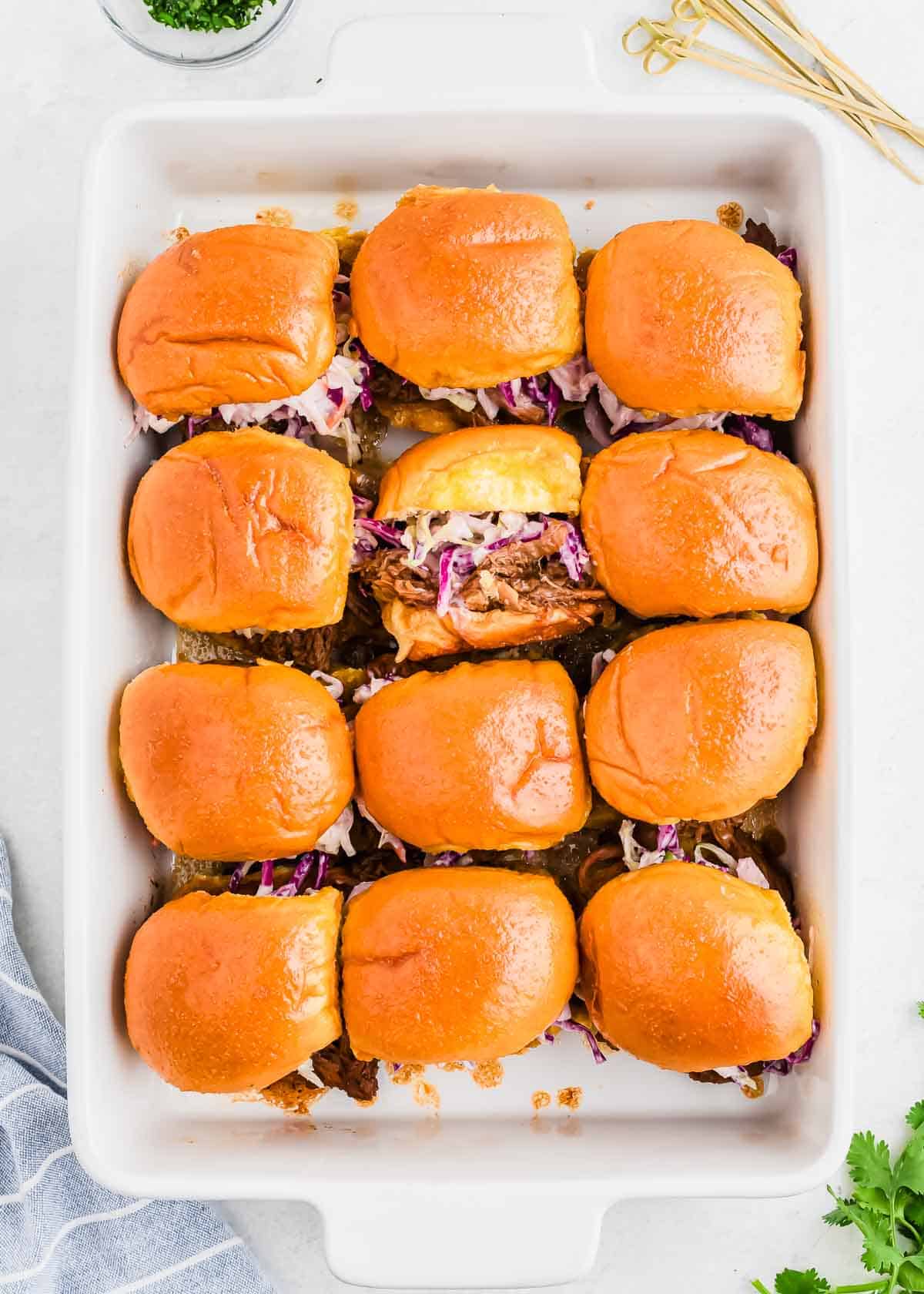 A baking dish filled with twelve pulled pork sliders, topped with coleslaw and placed on soft, golden-brown buns.