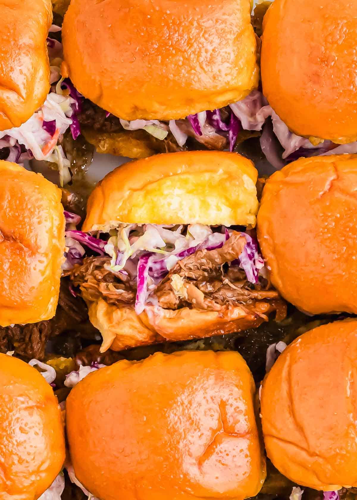 Close-up of several pulled pork sliders with coleslaw in soft buns, arranged closely together on a tray.