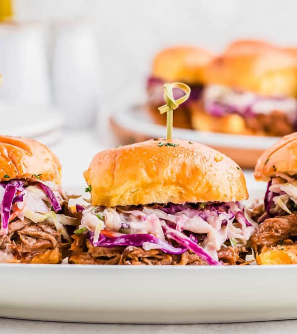 Three pulled pork sliders topped with coleslaw on a white serving plate, with another plate of sliders blurred in the background. Each slider is held together with a bamboo skewer.