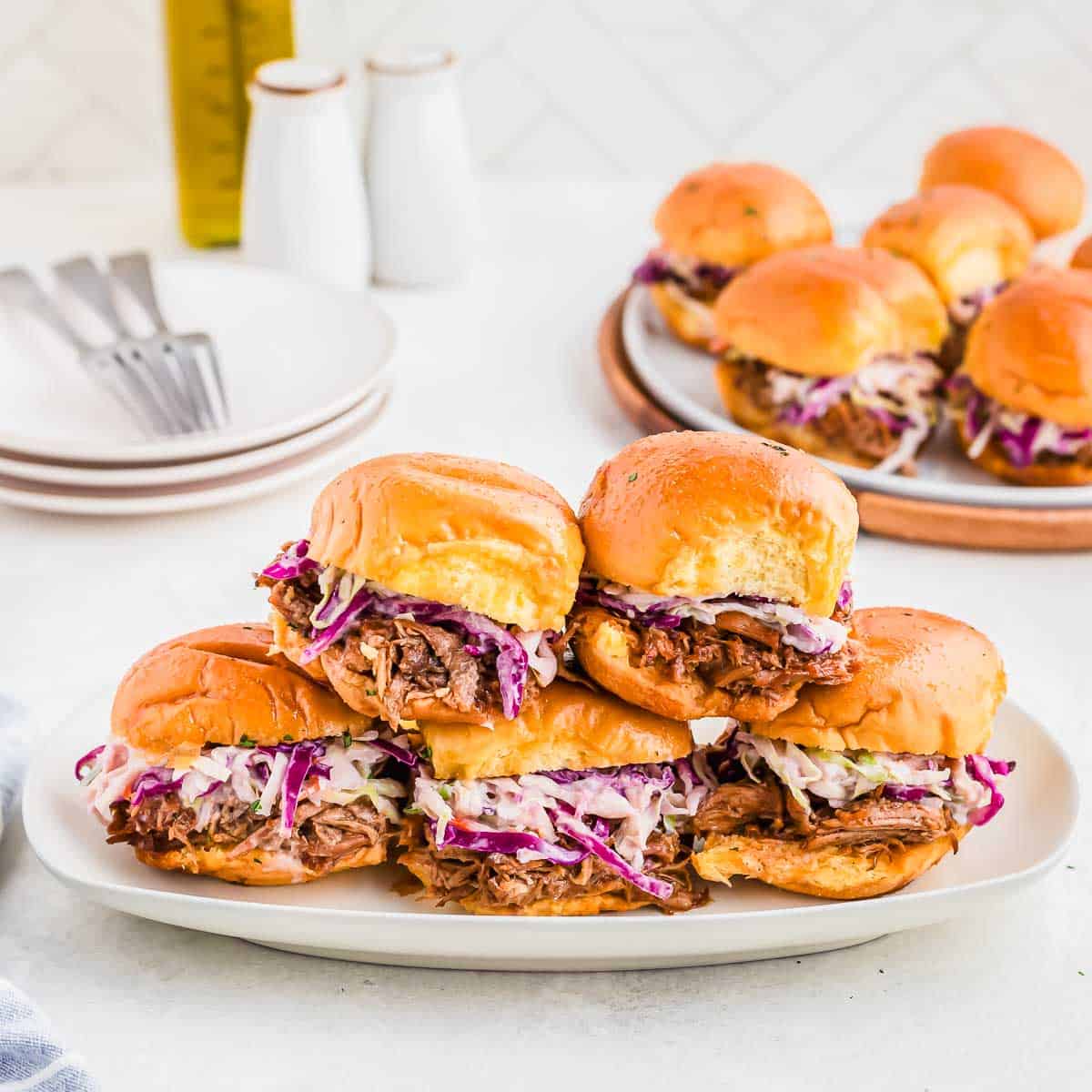 A plate of pulled pork sliders with coleslaw, with a set of forks, empty plates, salt and pepper shakers, and a tray of additional sliders in the background on a white surface.