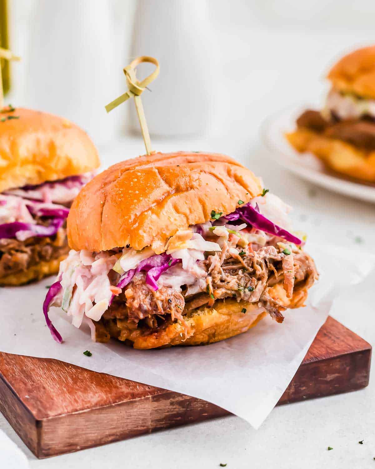 A pulled pork slider topped with coleslaw is served on a wooden board, with another sandwich visible in the background.
