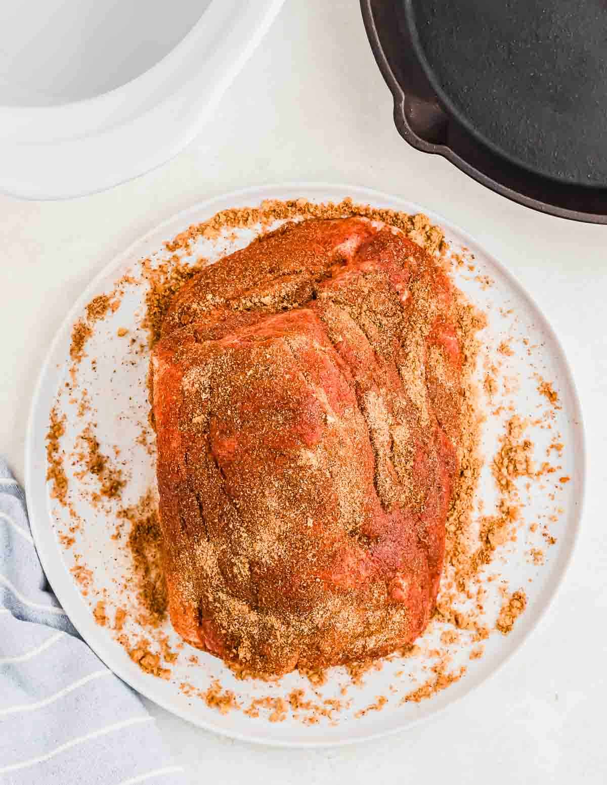 A piece of seasoned pork placed on a white plate, next to a black skillet and a white cloth on a white surface.