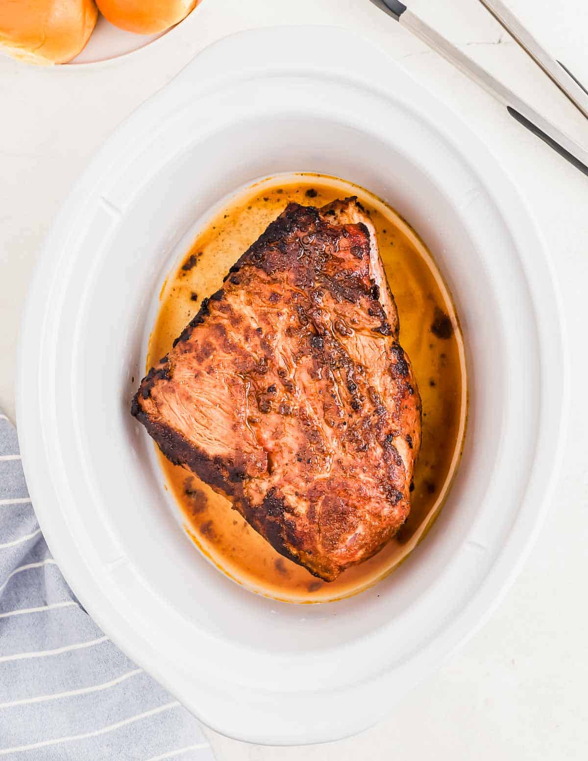 A cooked piece of pork in a white slow cooker, surrounded by juices. A pair of tongs is placed beside the slow cooker, with part of a striped cloth and oranges visible in the background.