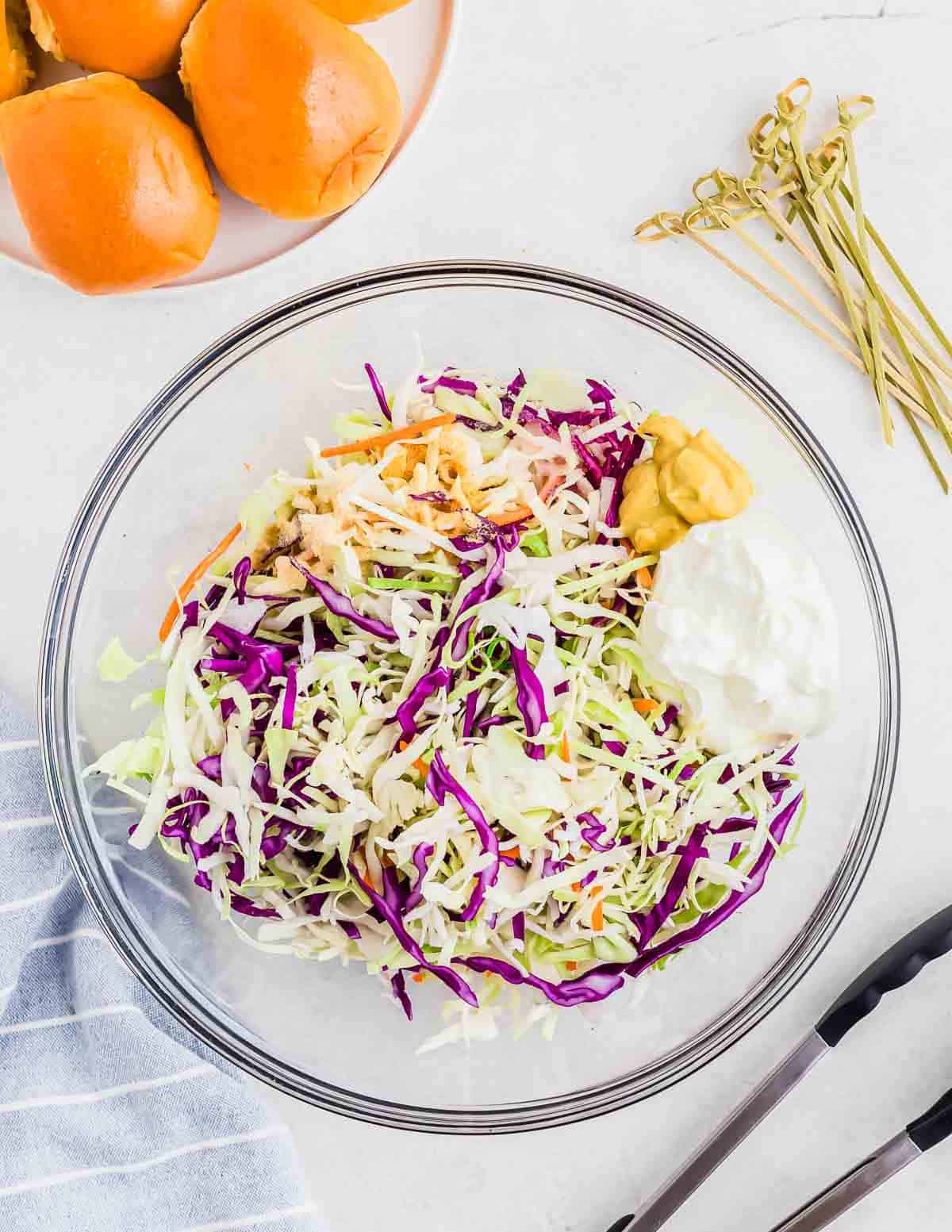 A clear glass bowl filled with shredded cabbage of various colors, a dollop of mustard, and yogurt, with dinner rolls and tongs on the side.