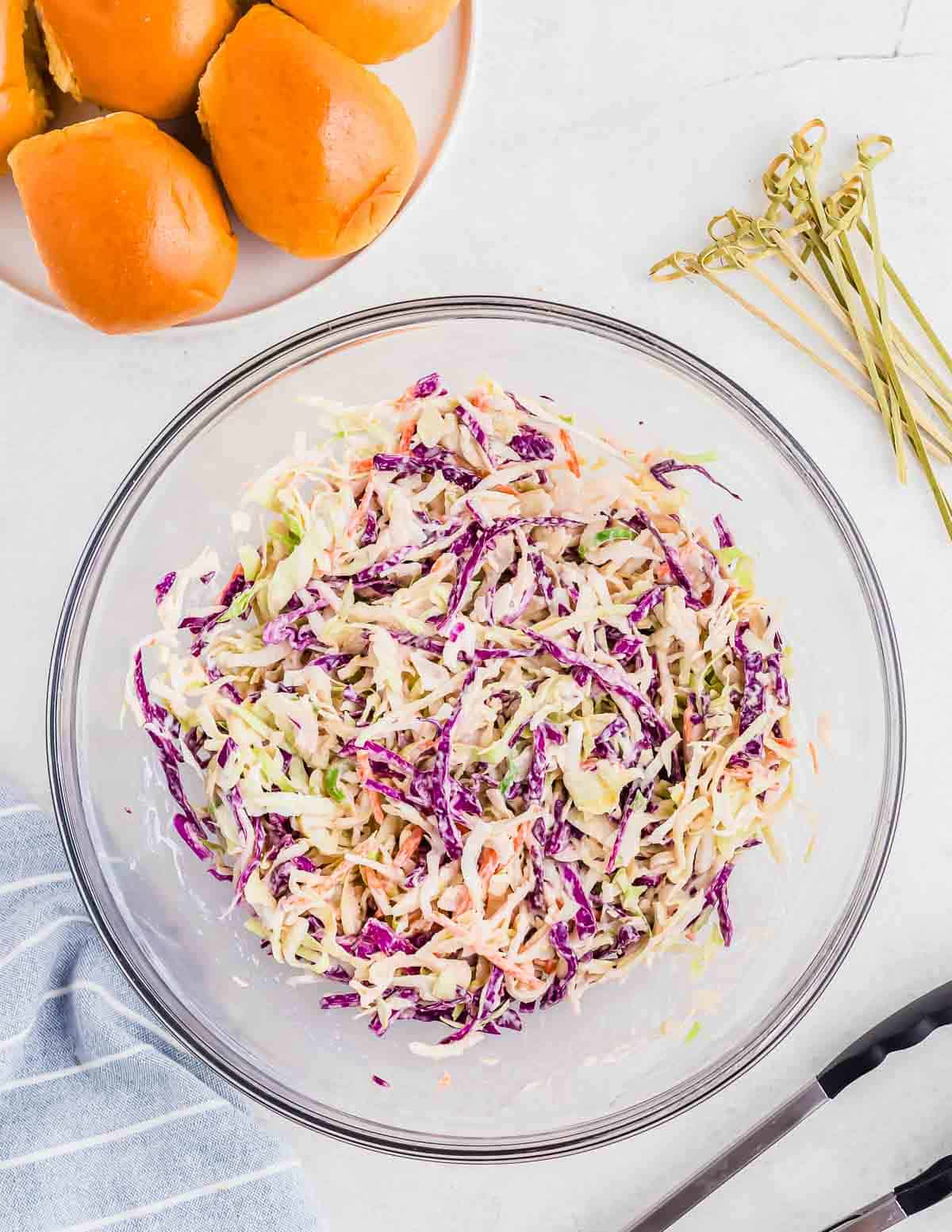 A glass bowl filled with coleslaw made of shredded cabbage and carrots, next to a plate of three dinner rolls and a striped napkin on a white surface.